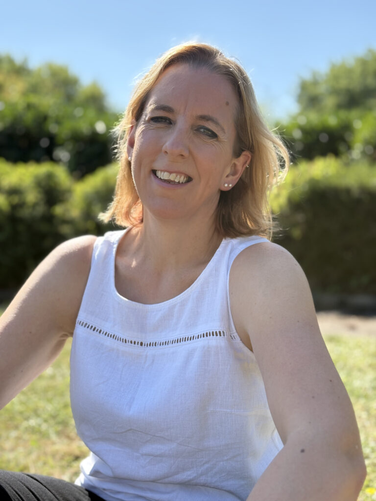 home. Photo of Fiona, The Habit Hacker, wearing a white top, sitting on grass in a park on a sunny day. Blue skies, trees and bushes in the background. Smiling and happy.