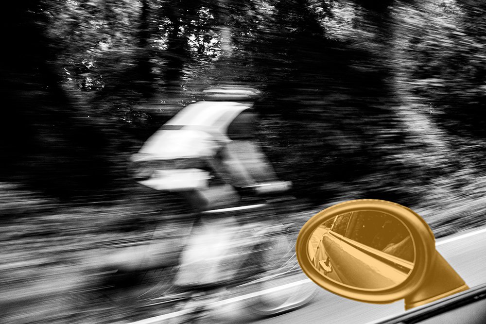 Black and white photo of a cyclist speeding past, shot from a moving car with a gold wing mirror in the foreground, representing forward momentum and questioning the road behind you.  