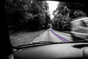 Black and white view from inside a moving car, blurred road with purple lines down the middle and trees through the windscreen, suggesting speed without clear direction. Busy going nowhere.