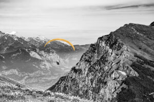 Black and white photograph of a paraglider soaring above a dramatic mountain landscape, valleys and peaks stretching into the distance below.