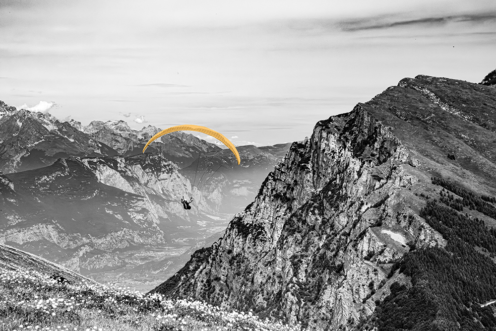 Black and white photograph of a paraglider soaring above a dramatic mountain landscape, valleys and peaks stretching into the distance below.  
