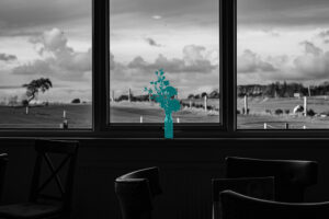 Black and white photograph looking through a cafe window at open countryside and dramatic cloudy sky, empty chairs in the foreground.