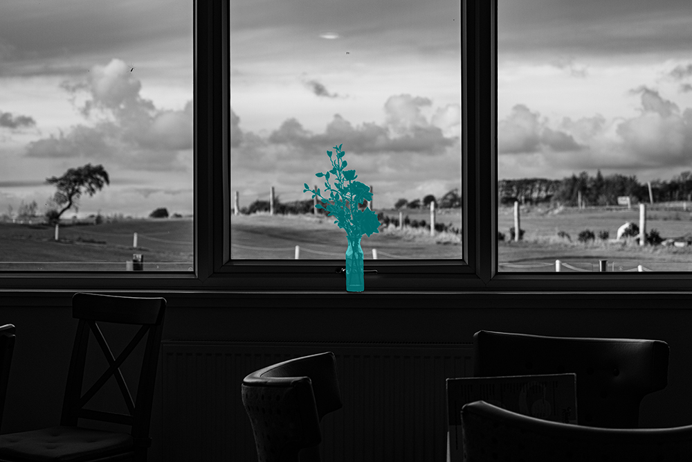 Black and white photograph looking through a cafe window at open countryside and dramatic cloudy sky, empty chairs in the foreground.  