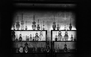 Black and white photograph of rows of glass bottles on backlit shelves, each a different shape and size, against a stone wall.