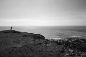 A person standing alone on a rocky headland, looking out across a calm sea in soft light contemplating why they feel like they’ve achieved nothing.
