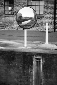 A convex security mirror on a brick warehouse wall, reflecting a wide view of a canal and surrounding buildings. Mirrors can help avoid solving the wrong problem.